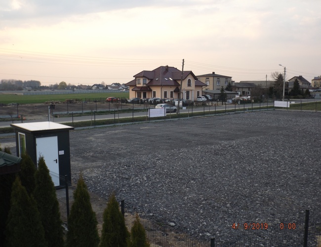 VIP Parking - Ogrodzony plac z nawierzchnią żwirową. W tle widoczne domy i pola. Na pierwszym planie mały budynek.