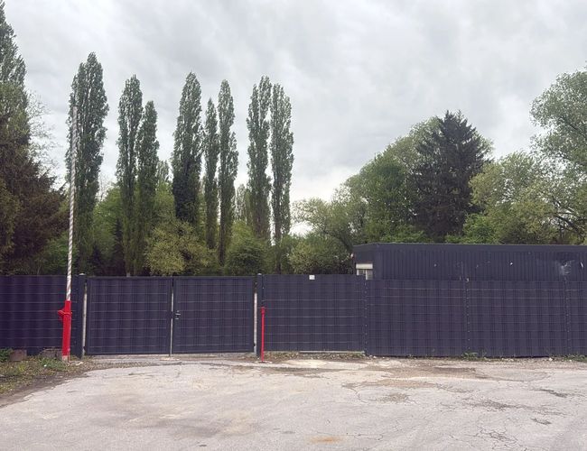 Parking Trust Park Fly - Ogrodzone miejsce z pobliskimi drzewami. Brama zamknięta, obok niebieski samochód.