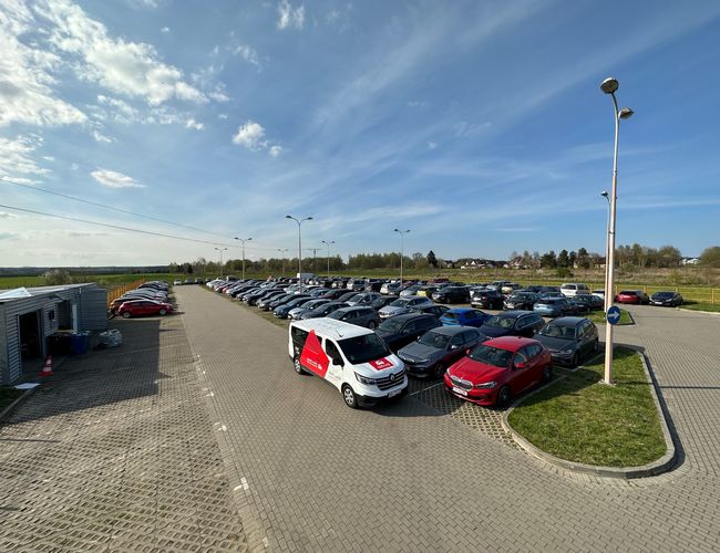 Odkryj-Parking GDN - Wiele samochodów stoi na szerokim parkingu. Wyraźne, wytyczone miejsca są wokół lamp ulicznych. Teren jest ogrodzony i zadbany.