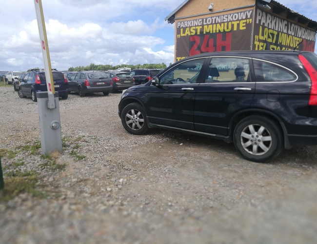 Parking P8 - Na parkingu stoją zaparkowane samochody. Teren jest nieutwardzony. W tle widoczny jest budynek z reklamą.