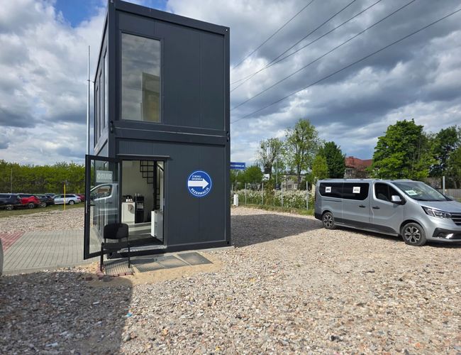 Parking Premium Parking – Na żwirowym terenie stoi szary kontener. Obok niego zaparkowano szary samochód. Obok widoczne są drzewa i kilka samochodów.