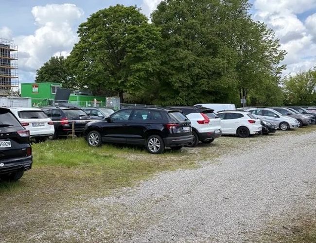 Parking terenowy z nawierzchnią żwirową i zaparkowanymi samochodami. W tle widoczne są wysokie drzewa i zabudowania.