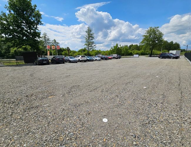 ParkZone - Na żwirowym parkingu stoi kilka samochodów. Teren otaczają drzewa i jest ogrodzony. Na niebie widać chmury.