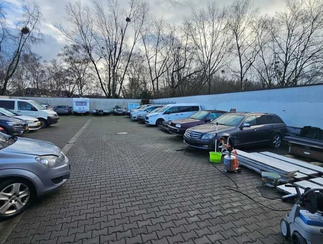 Parking Weltparking - Na placu stoją auta w dwóch rzędach. Teren jest utwardzony kostką, otoczony ogrodzeniem. Widać też drzewa.