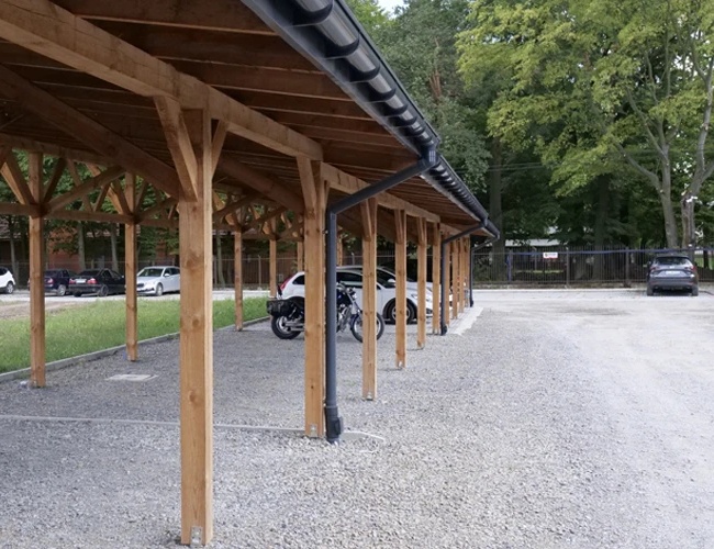 Parkovisko Zelený komplex Dwór Ostoya - Parkovisko má drevenú strechu. Pod jej strechou stoja autá a motorka. Na bočnej strane sú stromy.
