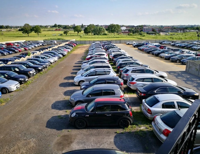 Parking Darex – En la imagen hay filas de coches aparcados. El área del parking está cercada y rodeada de vegetación. Cielo despejado de fondo.
