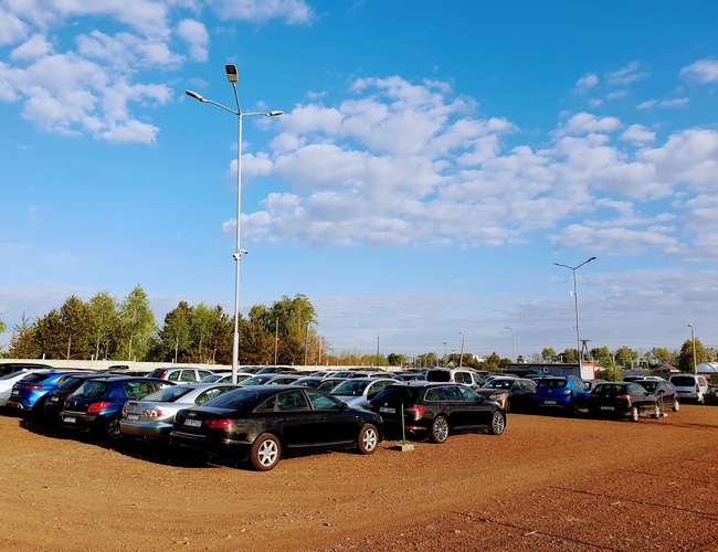 KTW Parking - Na parkingu stoją zaparkowane samochody. Jest teren nieutwardzony. Widać latarnie i zieleń w tle.