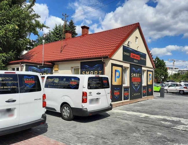 Smart Supreme Parking - Budova s červenou strechou a nápismi. Pred ňou je zaparkovaných niekoľko bielych vozidiel. Vedľa je vidieť aj ďalšie autá.
