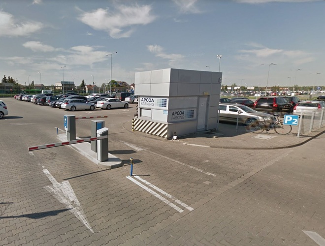 Aparcamiento oficial P2 – aparcamiento con entrada a través de una barrera. Al fondo muchos coches aparcados. El área está cercada y iluminada.