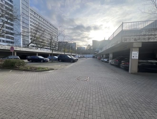 Valet Parking DUS - Parking jest przestronny i podzielony na strefy. Z jednej strony stoją zaparkowane samochody. Obok są budynki biurowe.