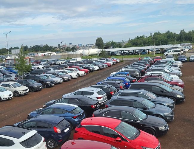 Sky Park – Na parkingu stoi wiele zaparkowanych samochodów. Teren jest otwarty, ale nieogrodzony. W tle znajdują się drzewa.