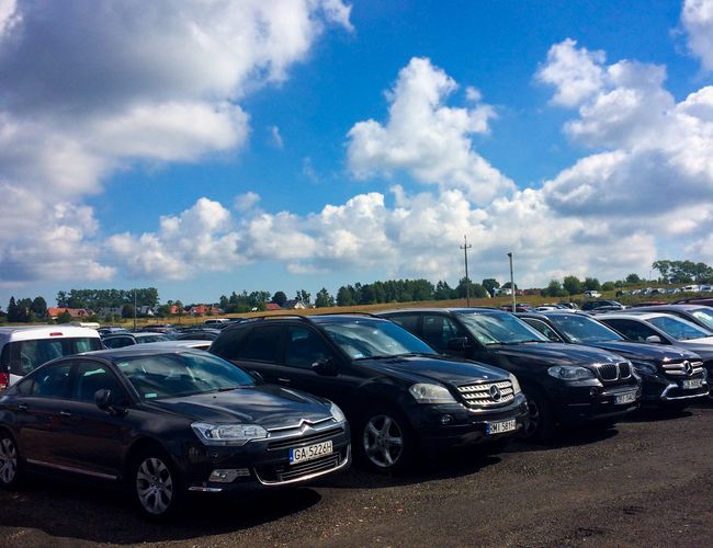 Parking Mega Park Service - Na placu stoi wiele samochodów osobowych. Jest słoneczny dzień z lekkim zachmurzeniem. Teren nie jest ogrodzony.