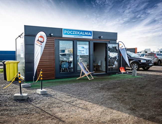 Parkovisko Okęcie Južné - pred čakárňou stoja zaparkované autá. Nachádzajú sa tam stoličky a lehátko. Pozemok je štrkový.