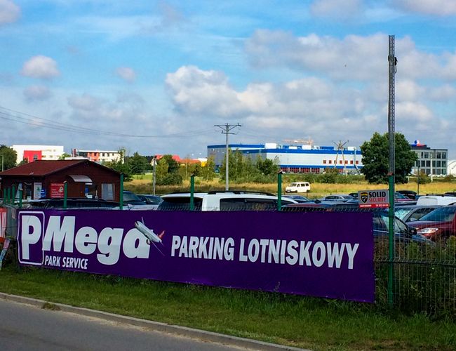 Parking Mega Park Service – Teren ogrodzony z widocznymi samochodami. Obok biegnie asfaltowa droga. W tle widać budynki i drzewa.