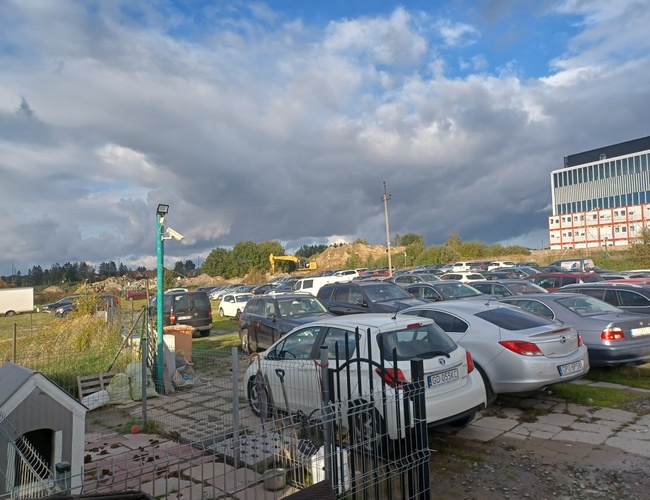 Maxi Parking - El área está llena de coches aparcados. También se puede ver una lámpara y una cámara. En el fondo se ve un gran edificio.