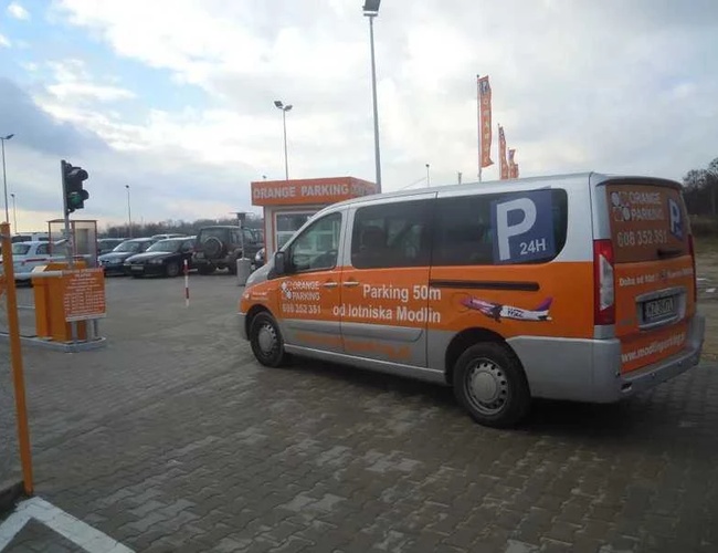 Orange Parking Modlin - Na parkovisku stojí autobus s reklamou. V pozadí sú viditeľné zaparkované autá a stánok. Chodník je dobre osvetlený.