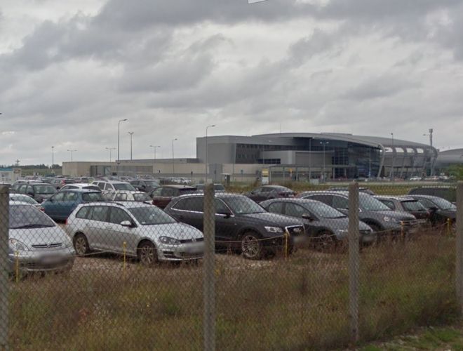Oficjalny Parking P6 – Parking jest ogrodzony siatką. Teren wypełniony samochodami. W tle znajduje się nowoczesny budynek.