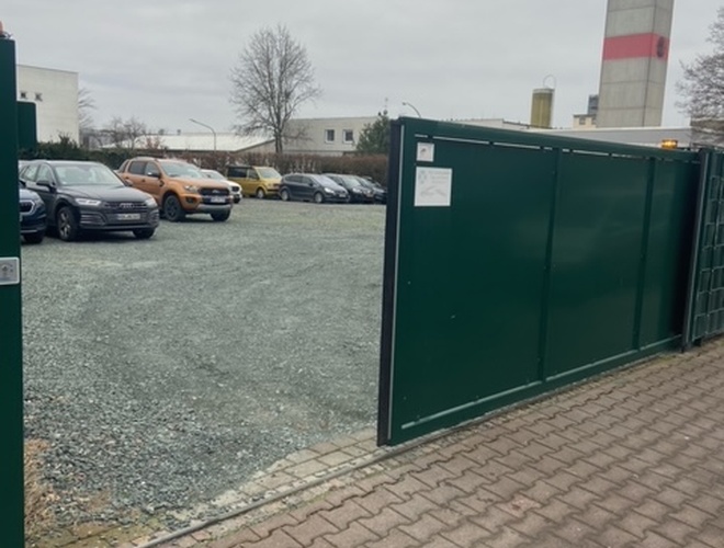Parkhaus Park-Box-Frankfurt Valet – Das Gelände ist eingezäunt. Geöffnete Einfahrtsgates. Sichtbare geparkte Autos, hauptsächlich im Hintergrund.