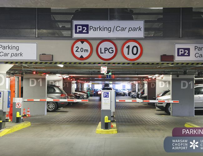 Oficjalny Parking Lotniska P2 – Kryty parking z wyraźnymi oznaczeniami przy wjeździe. Liczne samochody zaparkowane w rzędach. Barierki blokują wjazd.