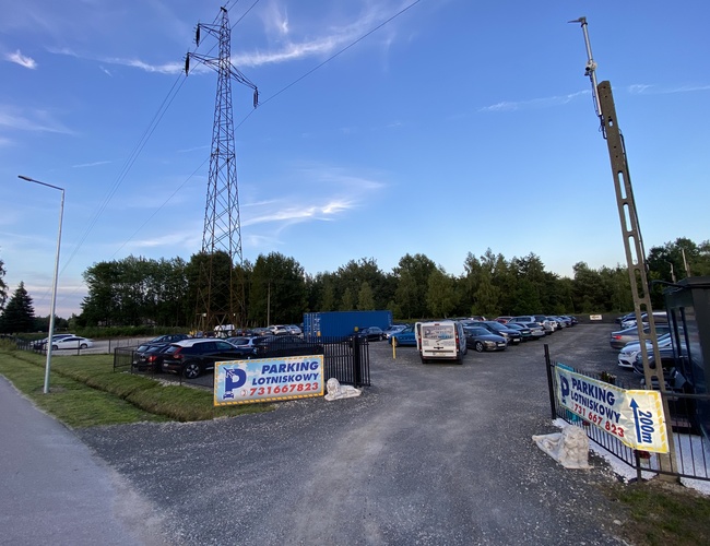 Parkovisková zóna výstupu - pri trase S7, ohradená s množstvom miest. Na pozadí sú vidieť stromy a energetická veža. Na parkovisku stoja osobné automobily.