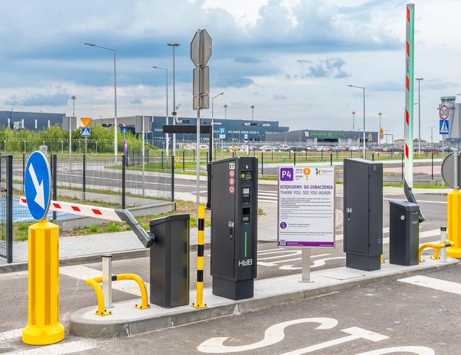 Aparcamiento Oficial P4 - Barrera de entrada y señales de tráfico. El área está cercada y iluminada. En el fondo se puede ver el edificio del aeropuerto.