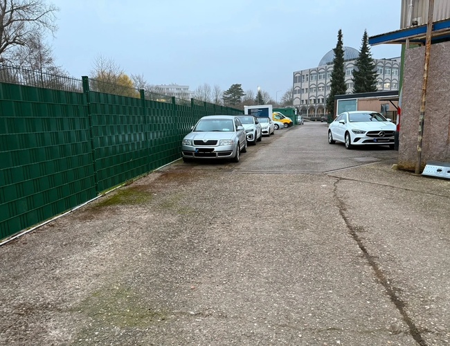 Parking Park Elite Valet - Samochody stoją wzdłuż zielonego ogrodzenia. Teren jest betonowy, prowadzi do budynku. W tle widać drzewa.