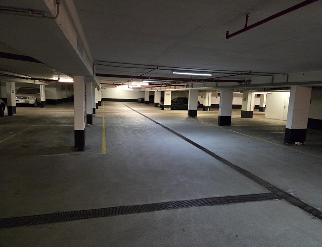 Atomis Hotel Munich Airport Parking Cubierto – Garaje subterráneo con suelo de hormigón. Se ven pocos coches y espacios señalizados. Hay pilares y tuberías en el techo.