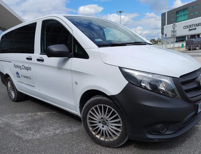 Parking Chopin - Biały minibus stoi na parkingu. Obok jest nowoczesny budynek hotelowy. Na niebie są białe chmury.
