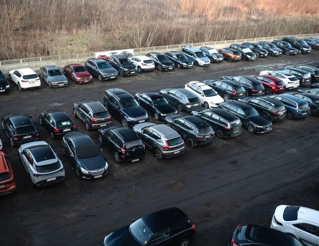 Auto Park Okęcie – Parking z wieloma samochodami. Teren ogrodzony betonowym płotem. W tle roślinność.