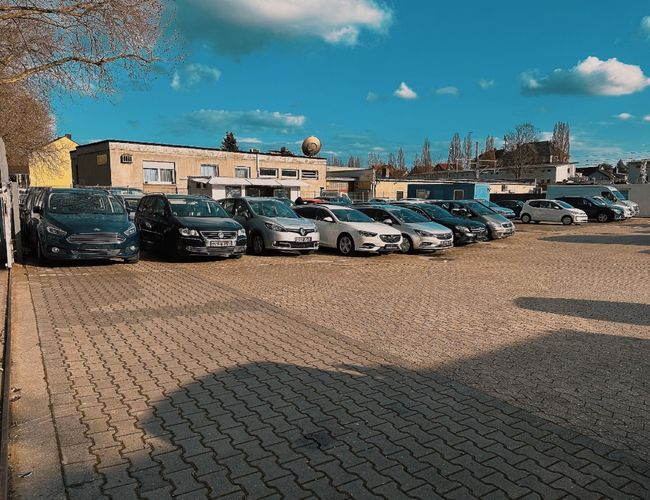 Parking Safetyparking24 VALET - Kilkanaście samochodów stoi na utwardzonej nawierzchni. W tle widoczne są budynki i drzewa.
