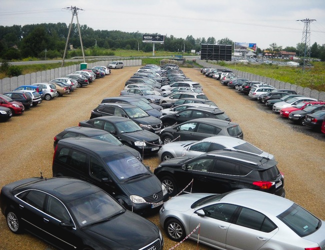 APARCAMIENTO DE VIAJES - El terreno está cercado y es de grava. Hay numerosos coches estacionados, colocados en paralelo. Al fondo se puede ver un área verde.