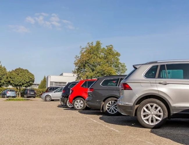 Parking Letim Brno II – na zdjęciu kilka zaparkowanych samochodów. Teren jest otoczony drzewami i budynkiem. Pogodny, słoneczny dzień.