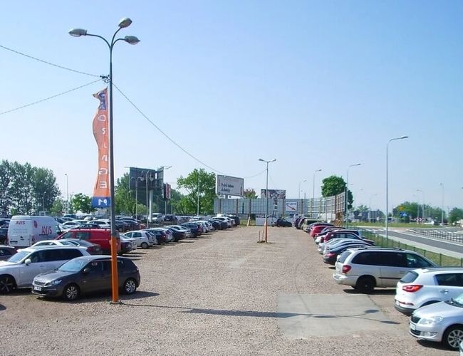 Orange Parking P3 – Parking ma utwardzoną powierzchnię. Jest ogrodzony siatką i dobrze oświetlony. Po bokach stoją zaparkowane samochody.