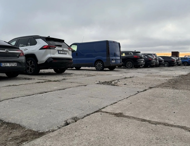 Parking MB Park – Na parkingu stoją auta osobowe i van. Podłoże jest betonowe i nierówne. Niebo jest pochmurne.