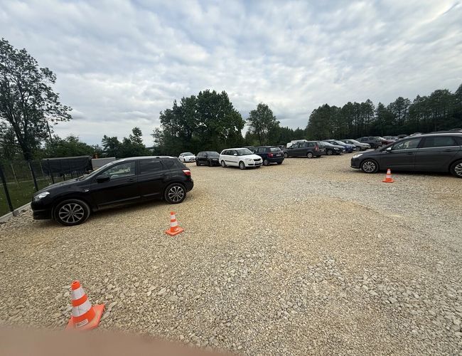 FlyParking – Parking żwirowy z wieloma samochodami. Teren otoczony drzewami. Rozstawione są pomarańczowe pachołki.