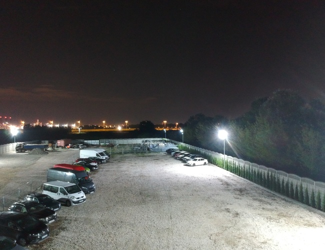 Aparcamiento del Aeropuerto 500 - El aparcamiento está iluminado y lo acompaña el cielo nocturno. Los coches están en filas sobre una superficie de grava.