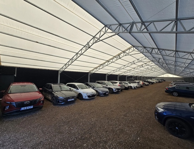 AleParking kryté - Vysoká zastrešená strecha zakrýva autá na štrkovom dvore. Vozidlá stoja vo rovnakých radoch po bokoch.