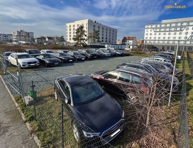 Parking Mein Valet Outdoor – Na zdjęciu widoczne są zaparkowane samochody. Teren jest ogrodzony. W tle znajdują się budynki.