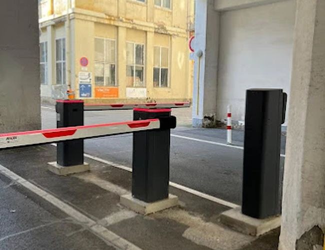 Parking Parkfinder24 Valet – Wjazd z szlabanem i słupkami. Teren zadaszony, otoczony jest budynkami. W tle widoczna jest żółta kamienica.