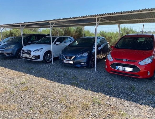 The Best Parking Alicante Covered Valet – Kilka samochodów stoi pod wiatą. Teren parkingu jest ogrodzony siatką. W tle widać krzewy.