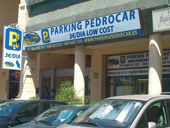 Aparcamiento PedroCar – Se puede ver un edificio con un gran cartel. Delante del edificio hay coches aparcados. Parte de las ventanas están acristaladas.