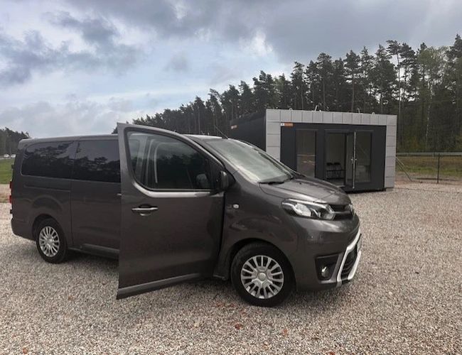 Auto Airport Parking - Dostawczy van z otwartymi drzwiami stoi na żwirowym placu. Za nim widać niewielki budynek.