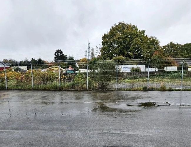 ParKing’s Düsseldorf Outdoor Valet – mokry betonowy plac za ogrodzeniem. Teren otoczony metalową siatką. W tle drzewa i kontenery.