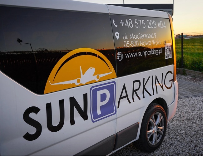 Parkovisko Sun - Biela dodávka s logom a kontaktnými údajmi. Stojí na kamienkovej povrchovej úprave pri oplotení zo sieťoviny. Na pozadí je vidieť západ slnka.