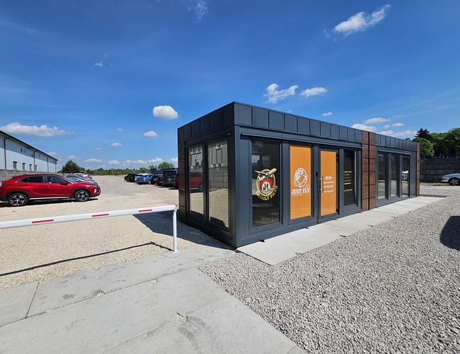 Just Fly Parking – nowoczesny budynek biurowy i zamknięty szlaban. Na parkingu stoi kilka samochodów osobowych. Obok znajduje się żwirowa nawierzchnia.