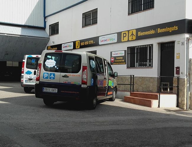 Lowcost Parking Walencja - Furgonetki stoją przy budynku z szyldem. Drzwi są otwarte. W tle widoczna hala magazynowa.