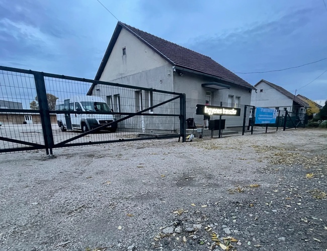 Brama wjazdowa na żwirowy plac prowadzący do budynku przy parkingu, otwarta przestrzeń i pochmurne niebo.