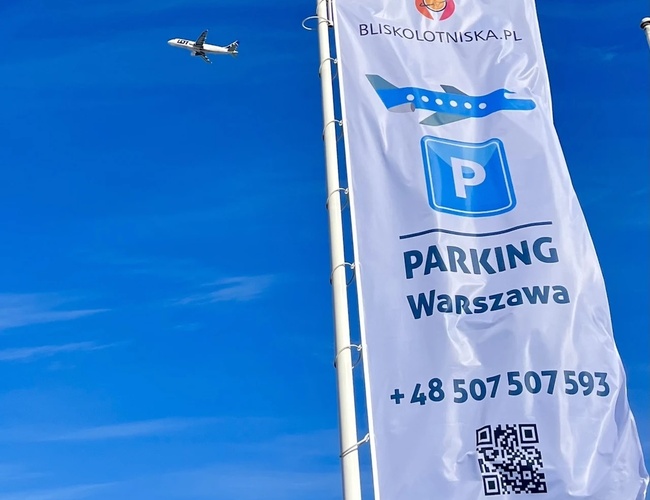 Parkovanie blízko letiska - Biela vlajka s nápisom Parking Warszawa. Na oblohe je vidieť lietajúce lietadlo.