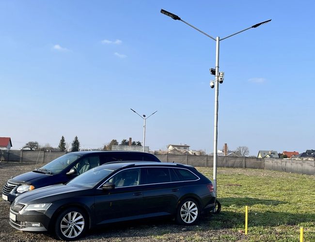 Parking CARPORT RDO - Na placu stoi latarnia z kamerami. Obok są dwa zaparkowane samochody, jeden czarny i jeden granatowy.
