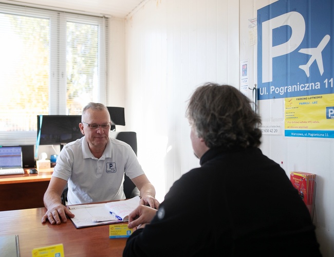 Parkuj Leć – Dwie osoby rozmawiają przy biurku w biurze. Na ścianie widnieje logo parkingu.